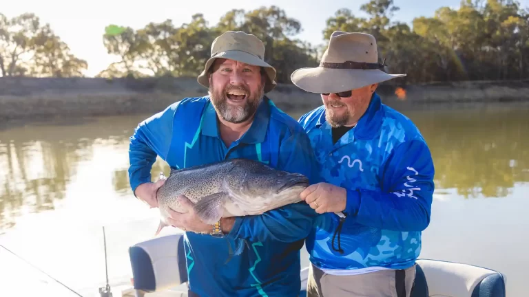 Two excited fishermen in blue shirts and hats proudly displaying large Murray cod catch on a boat in Tropical North Victoria